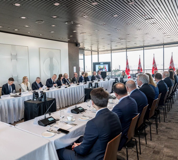 Des membres du Comité consultatif sur les relations économiques entre le Canada et les États-Unis sont assis autour d’une grande table de conférence lors d’une réunion, avec des microphones, des cartons nominatifs et des drapeaux du Canada visibles dans la salle.