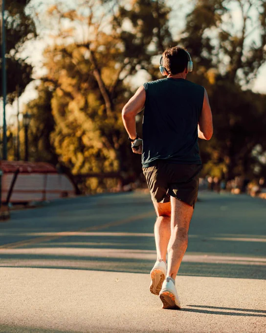 A person is jogging on a sunny road lined with trees, wearing headphones, a sleeveless shirt, and shorts. The scene is calm and peaceful.