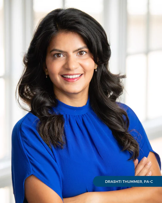 Drashti Thummer, PA-C, of Tryon Medical Partners wears a blue shirt and poses with her arms crossed in a professional portrait in front of a paned glass window