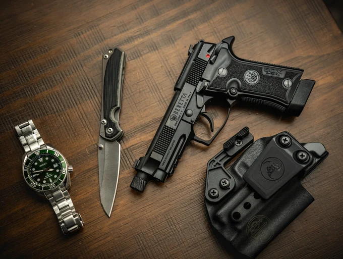 A silver watch with a green face, a folding knife, a Beretta handgun, and a black holster are arranged on a wooden surface.