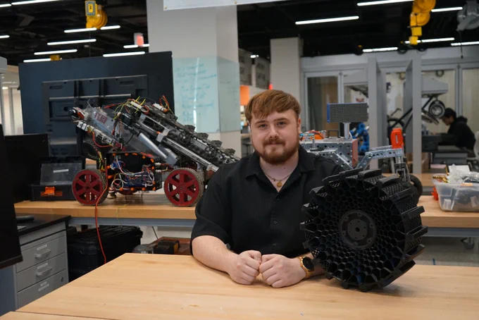 A student is sitting at a table in a robotics lab, surrounded by mechanical equipment and a complex robot on the left. A prototype of the wheel is next to the student on the table.