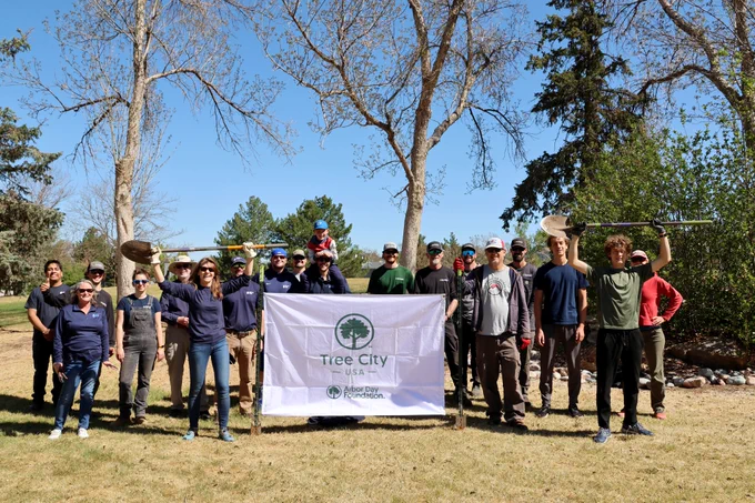 Group of people standing together on a grass lawn next to a white flag that says, "Tree City USA. Arbor Day Foundation."