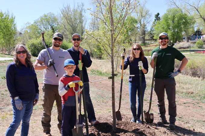 Group of three men, two women, and young boy standing together, smiling next to a freshly planted tree.