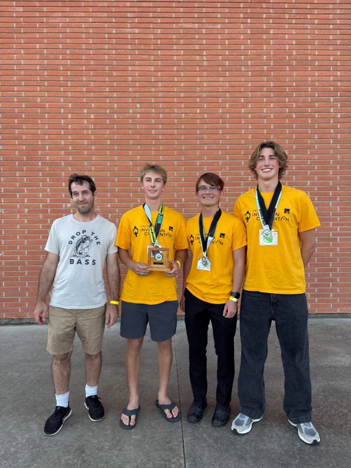 Four young men standing in front of a brick wall, three wearing yellow shirts with medals and one holding a trophy, celebrating a competition win.