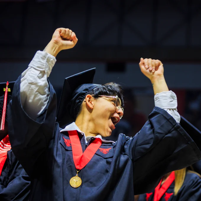 A graduate in a cap and gown raises both fists in celebration during a commencement ceremony, wearing a gold medal and red honor cord.