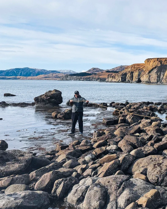 Person standing on rocky shore holding a large seaweed with cliffs and calm sea in the background under a partly cloudy sky.