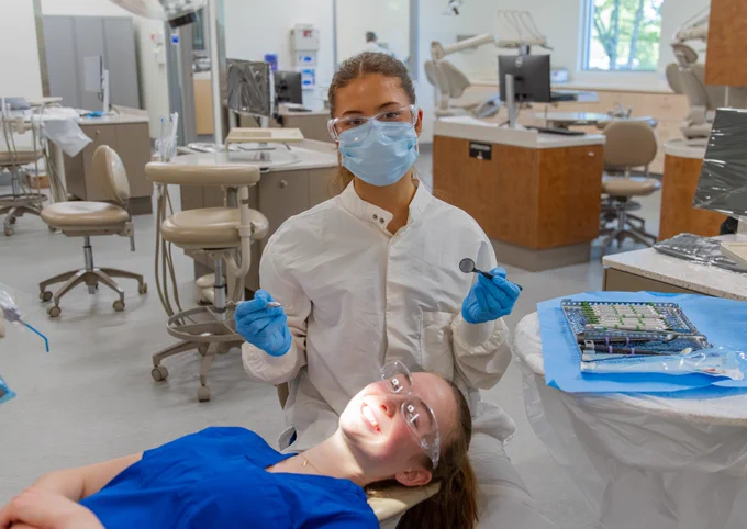 A dental student wearing a mask and gloves is holding dental instruments in a clinic. A student patient is reclining in the dental chair, wearing protective glasses. They're both smiling at the camera. The room has dental equipment and workstations.