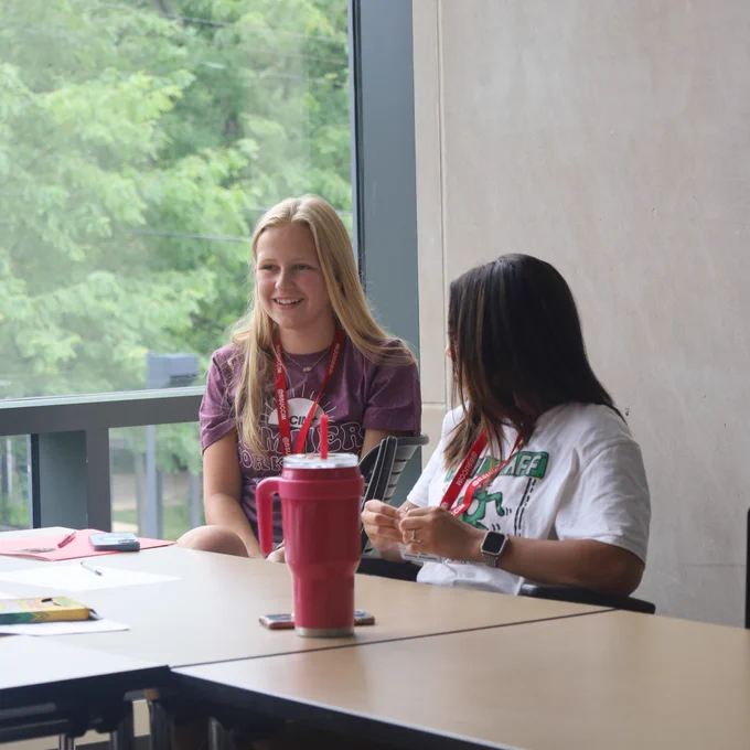 Two students wearing lanyards sit together at a table near a large window, engaged in conversation, with a water tumbler and papers spread across the table.