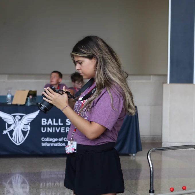A student reviews images on a Canon DSLR camera while standing in a lobby in front of a Ball State University College of Communication, Information, and Media table.