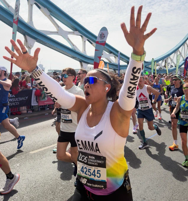 Runners cross Tower Bridge during in the London Marathon. In the foreground, a woman named Emma raises her arms and cheers as she runs, surrounded by fellow marathoners.