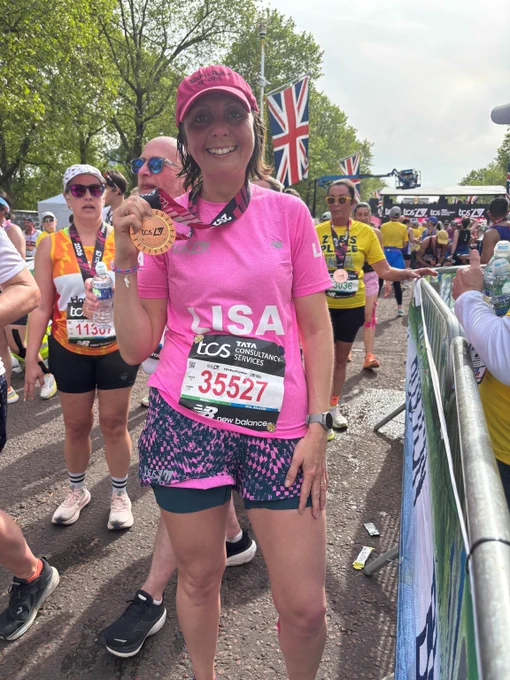 Runner in pink shirt and patterned shorts holding a medal after completing a marathon event with Union Jack flags in the background.