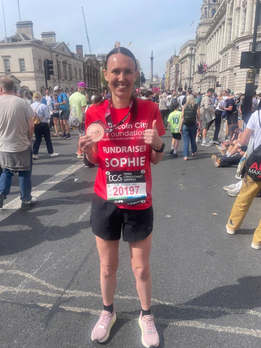Runner Sophie in a red Lincoln City Foundation shirt holds a medal after completing a marathon in a crowded city street.