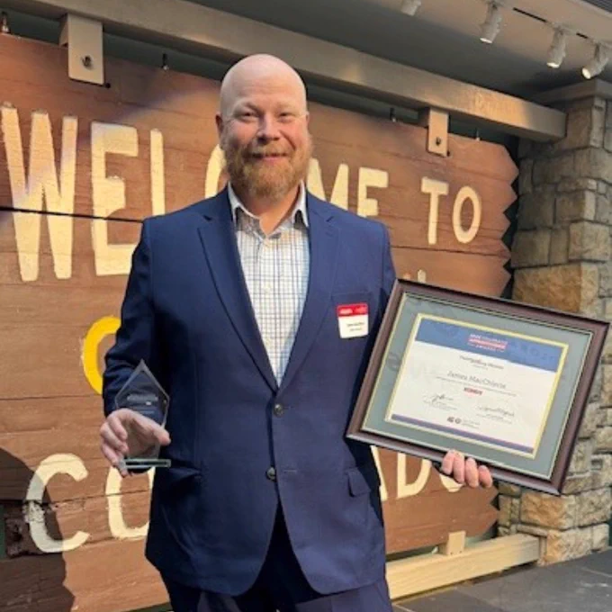 Man in navy suit holding a framed certificate and a glass award standing in front of a wooden Welcome to Colorado sign.