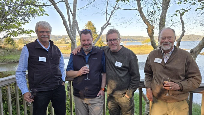 Four men standing on a wooden deck overlooking a river, each holding a glass of wine.