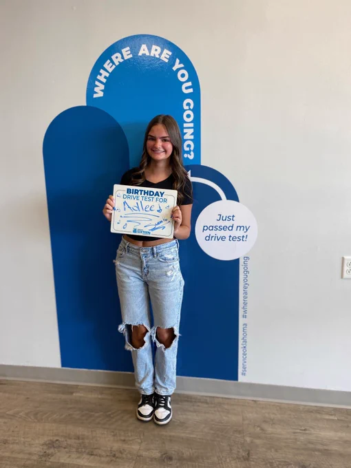 A person stands smiling, holding a "Birthday Drive Test for Adlee" sign. They are in front of a wall with a blue design featuring the phrases "Where are you going?" and "Just passed my drive test!" The person is wearing ripped jeans and standing on a wood floor.