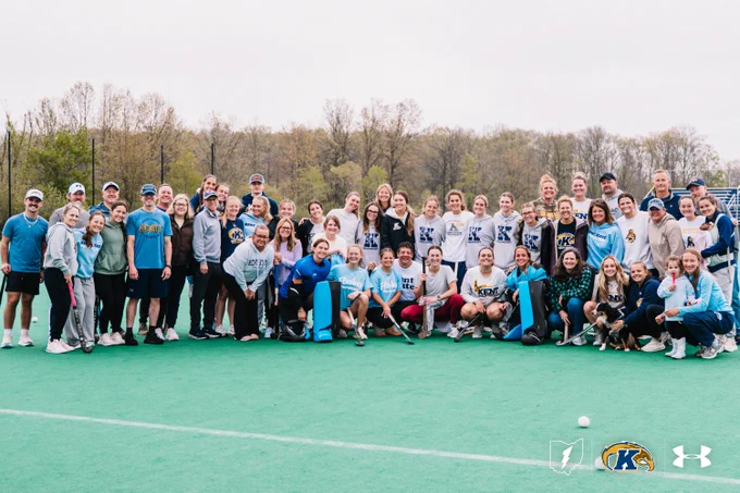 A large group of people posing together on a sports field, wearing athletic and casual clothing. The sky is overcast, and trees are visible in the background. Several individuals are wearing shirts featuring the Kent State logo. Sports equipment and logos from Under Armour and Kent State are visible on the field.