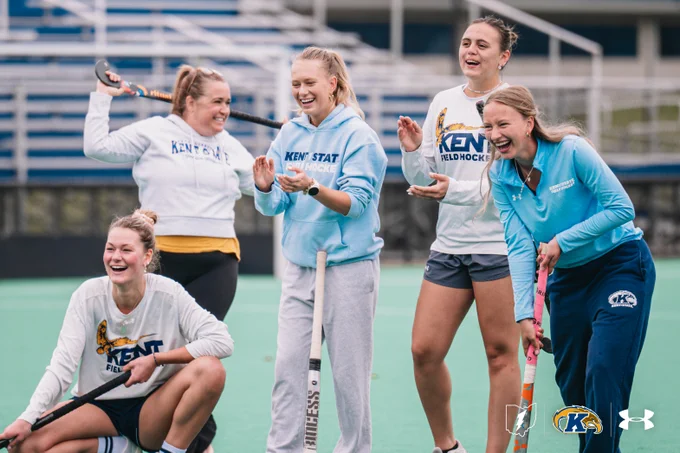 A group of five people are happily gathered on a sports field. They are wearing Kent State field hockey attire. Some are holding hockey sticks, and the field is lined in green with a blurred seating area in the background. The Kent State and Under Armour logos are visible in the image.
