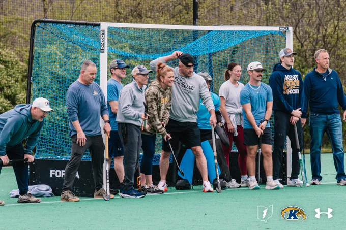 A group of people are lined up on a sports field, holding hockey sticks in front of a goal net. Some are wearing athletic attire and caps, while others are in casual clothing. Logos for Kent State and Under Armour are visible on the bottom of the image.