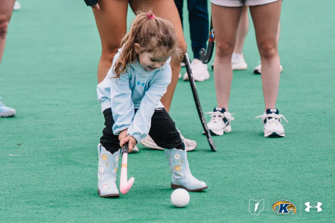 A young child on a playing field is preparing to hit a ball with a hockey stick. Several adults stand nearby, holding sticks. The child is wearing blue boots with cartoon designs. Logos for Kent State, Under Armour, and another entity are at the bottom of the image.
