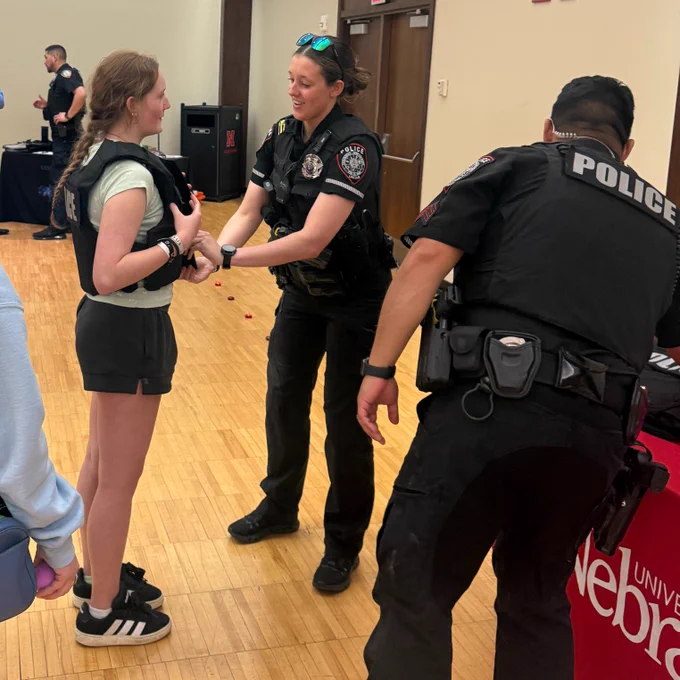 A police officer demonstrates how to wear a protective vest for a student at a criminal justice career fair, while other attendees watch and interact with officers at a University of Nebraska booth.