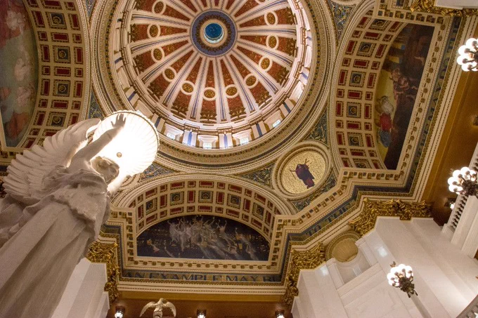 A picture from the ground level looking up at an ornate interior with a domed ceiling, intricate murals, and gold detailing. A statue of an angel with wings is in the foreground, lit warmly. Photo courtesy of https://www.reverberations.net/pennsylvania-state-capitol-tour/