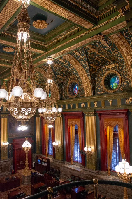 A photo of an ornate, historic hall with intricately designed ceiling featuring gold accents, grand chandeliers, stained glass, and luxurious drapes on the windows. On the carpeted floor are wooden desks and more ornate lamps. Photo courtesy of https://www.reverberations.net/pennsylvania-state-capitol-tour/