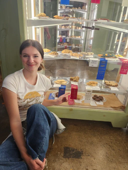 A young girl with brown hair wearing blue jeans and a tan shirt with horses on it, pointing to a plate of cookies inside a glass case. The plate has a red, second-place ribbon hanging from it. 