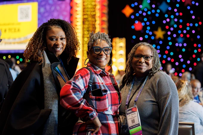 Photo of three people standing, posing, and smiling at the camera. Behind them, there are many other people sitting and standing around tables. There is a big presentation board and colorful, glowing lights decorating the presentation hall. 