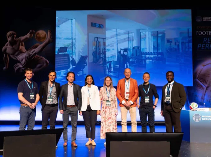 A group of eight people stand side by side on a stage with a large screen in the background displaying a gym. They appear professional and are smiling.