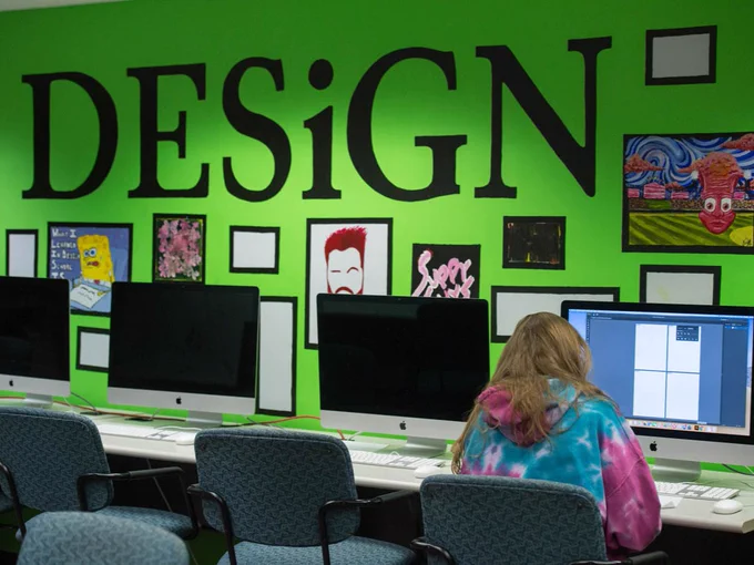 A person in a colorful hoodie is sitting at a computer in a design classroom. The wall is bright green with the word "DESIGN" in large black letters surrounded by framed artwork. Several computers are lined up on the desk.