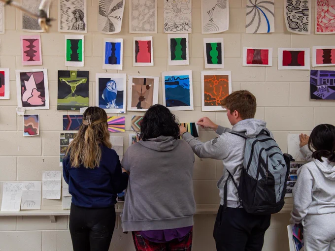 A group of people stand in an art classroom, viewing and adjusting colorful artwork displayed on a wall. They are examining various abstract and patterned pieces, with more artwork scattered on a nearby table.