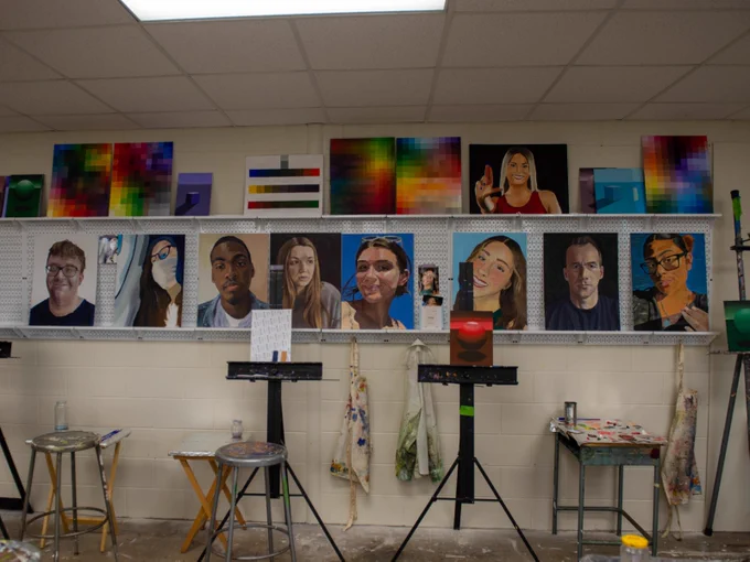 This image depicts an art classroom displaying various portrait paintings on shelves and easels. The paintings vary in style and include different individuals. The background also features abstract color grids and palettes. The room is equipped with stools, tables, and art supplies.