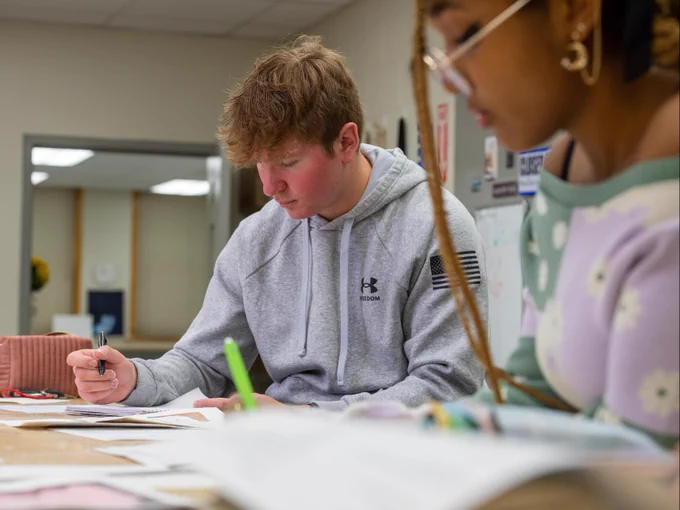 Two people are focused on studying at a table covered with books and papers. One person is wearing an Under Armour hoodie and writing on paper. The other is wearing glasses and a patterned sweater. The background shows a whiteboard and office decor.