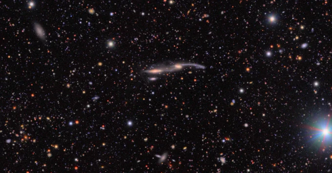 Two interacting galaxies at center, with cores and extended tendrils of stars in a thin horizontal line. The background is black, sprinkled with countless galaxies and stars of varying colors and sizes.