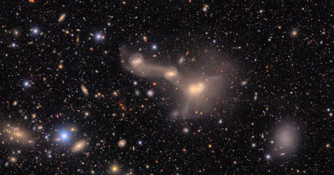 A complex entanglement of three interacting galaxies, bright cores lined up left to right and connectied by spiderweb-like tendrils. The background is black, sprinkled with countless galaxies and stars of varying colors and sizes.