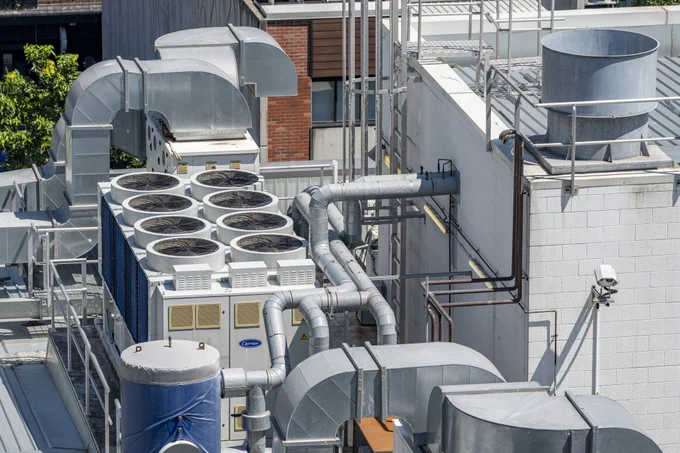 A rooftop with an industrial air conditioning system, including multiple large fans, ductwork, and vents. The equipment is marked with a Carrier label, surrounded by buildings and some greenery visible in the background.