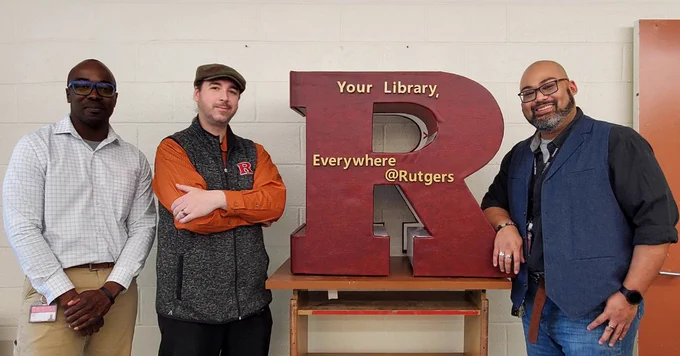 Three people stand in front of a large red letter "R" with the text "Your Library, Everywhere @Rutgers" on it. They are posing and smiling for the photo.