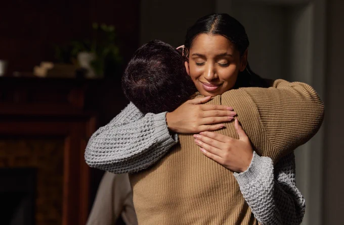 Two people embracing warmly indoors, one wearing a beige sweater and the other a gray knit sweater, conveying comfort and support.