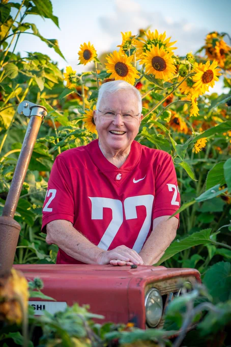 Regent Sam Hunt stands in a sunflower field, wearing a crimson WSU football jersey. Tall sunflowers and green foliage surround him, with yellow blooms visible against a bright sky in the background.
