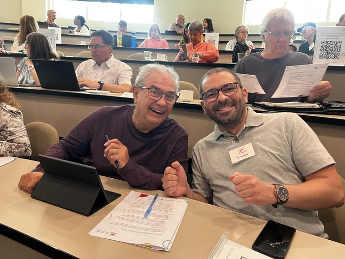 A Connected Canadians volunteer sitting with an older adult during a workshop, with other participants using devices in the background.