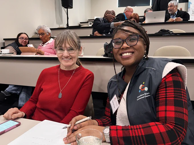 An older adult and a Connected Canadians volunteer smiling together during a digital skills workshop, with other participants working on devices in the background.