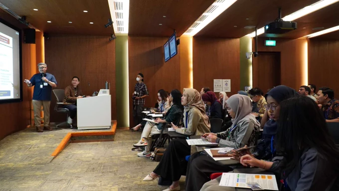 Presenter standing to the front of the room, presenting to participants seated in a modern conference room with wood-paneled walls.