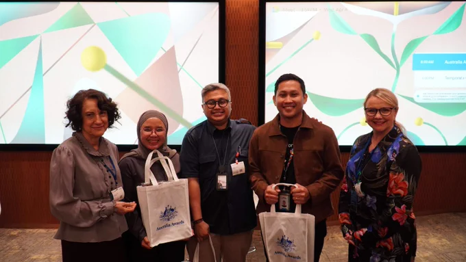 Photo of the panel standing indoors holding tote bags with a logo, posing in front of two large projector screens.