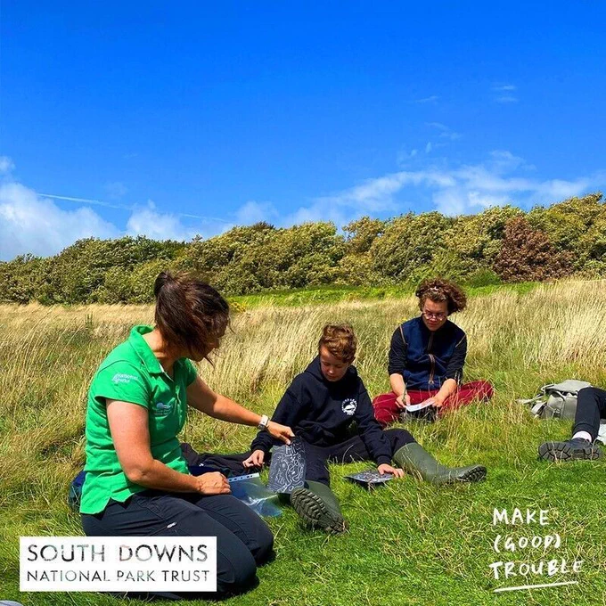 Group of children sitting in a field with long grass looking at art being shown by a SDNPA member of staff.