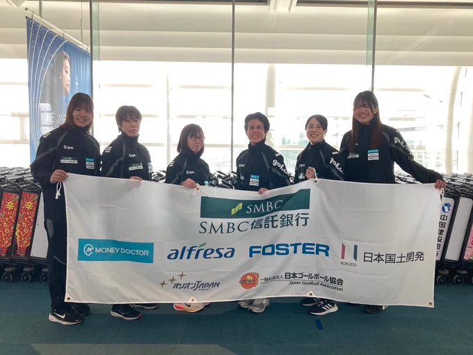 空港で日本代表女子の6人が横に並び、白地の横断幕を持って立っている。横断幕にはSMBC信託銀行、マネードクター、アルフレッサ、フォスター電機、日本国土開発のロゴ、オリオンJAPANとゴールボール協会のロゴが掲載されている