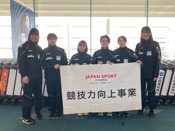 空港で日本代表女子チームが競技力向上の横断幕を持って横に6人並んで立つ様子