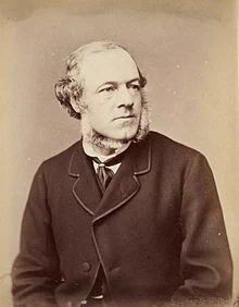 Black and white photograph of a man. He has grey-white hair and a grey-white beard. He is wearing a dark jacket and necktie.