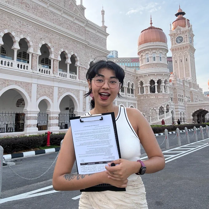A young Indian woman is standing, smiling with a petition document in hand. She is wearing a white top and peach skirt. She is standing in front of a peachy looking historical building, known as Sultan Abdul Samad building.