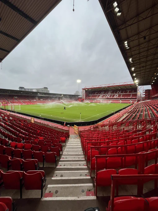 Photo taken from the back corner of the Bridgford stand at Nottingham Forest’s City Ground. A row of steps run down at the bottom of the image, flanked by empty red seats. In the middle of the shot is a green pitch which has a set of sprinklers watering it. The single tier Peter Taylor Main Stand is in the left whist the double tier Trent End can be seen at the top of the image. The photo was taken in daylight but a solitary floodlight is switched on in the top left of the image.