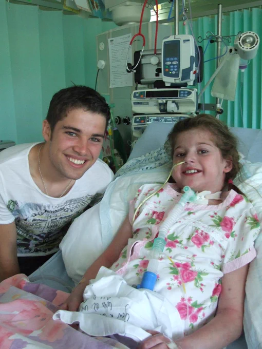 Smiling girl in hospital bed with tracheostomy and nasal feeding tube; a young man beside her; medical monitors behind.
